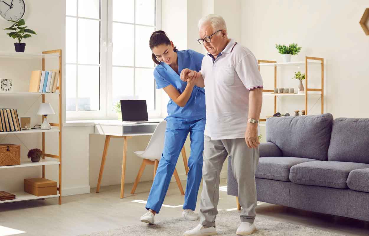 A senior living placement specialist assisting an elderly man off of the couch.